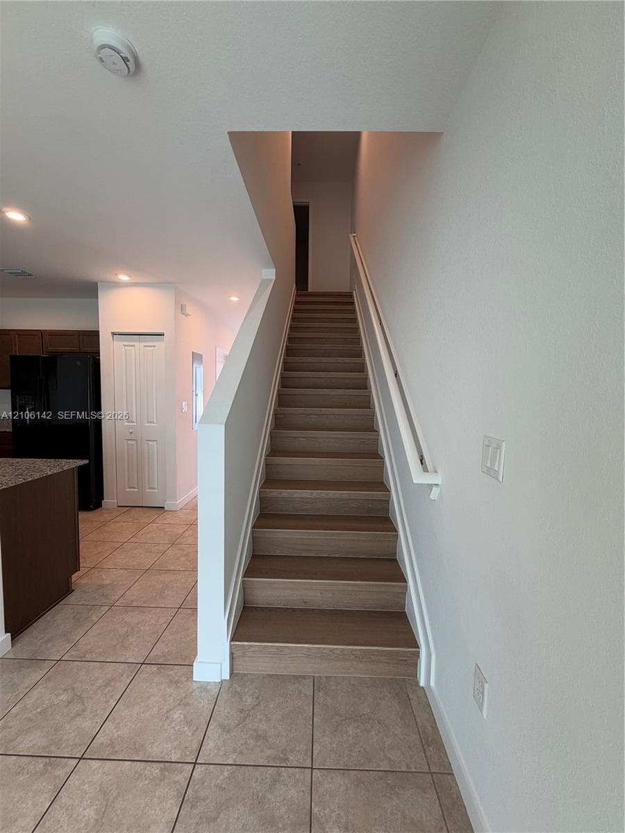 Staircase featuring wood-finish treads, a white handrail, and ceramic tile flooring throughout the main level
