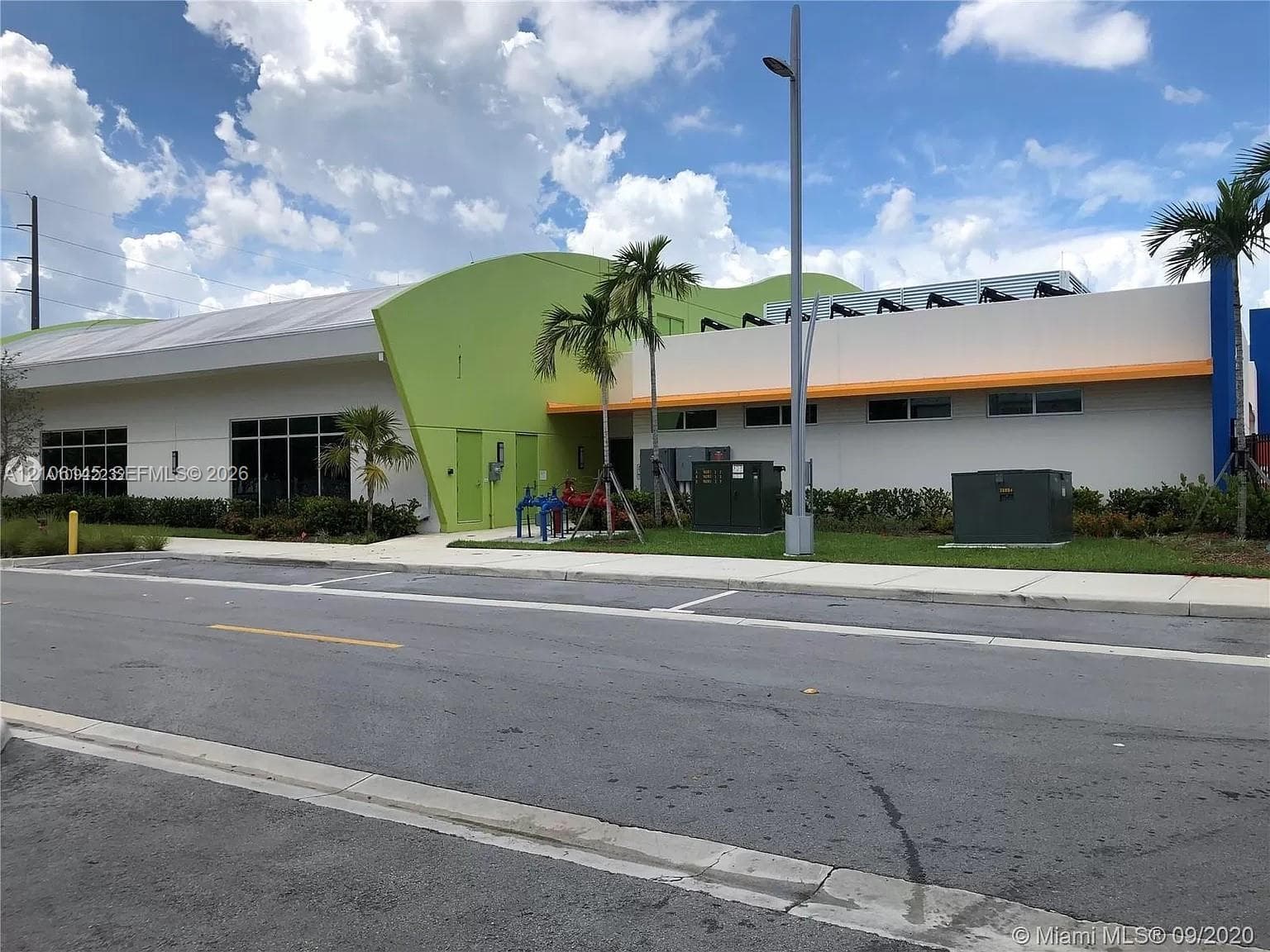 Modern commercial exterior featuring a curvilinear lime green architectural accent, concrete sidewalk, multi-pane glass windows, and palm tree landscaping