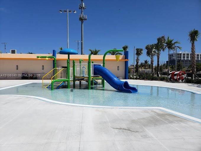 Splash pad featuring a central play structure, blue slide, overhead water features, and expansive concrete decking