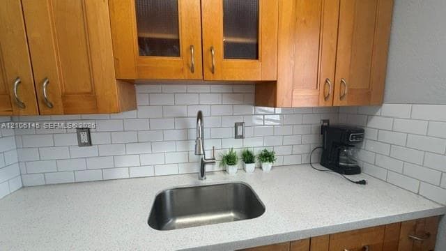 Kitchen featuring wood cabinetry with glass-panel inserts, white subway tile backsplash, light-toned composite countertop, undermount stainless steel sink, and high-arch pull-down faucet