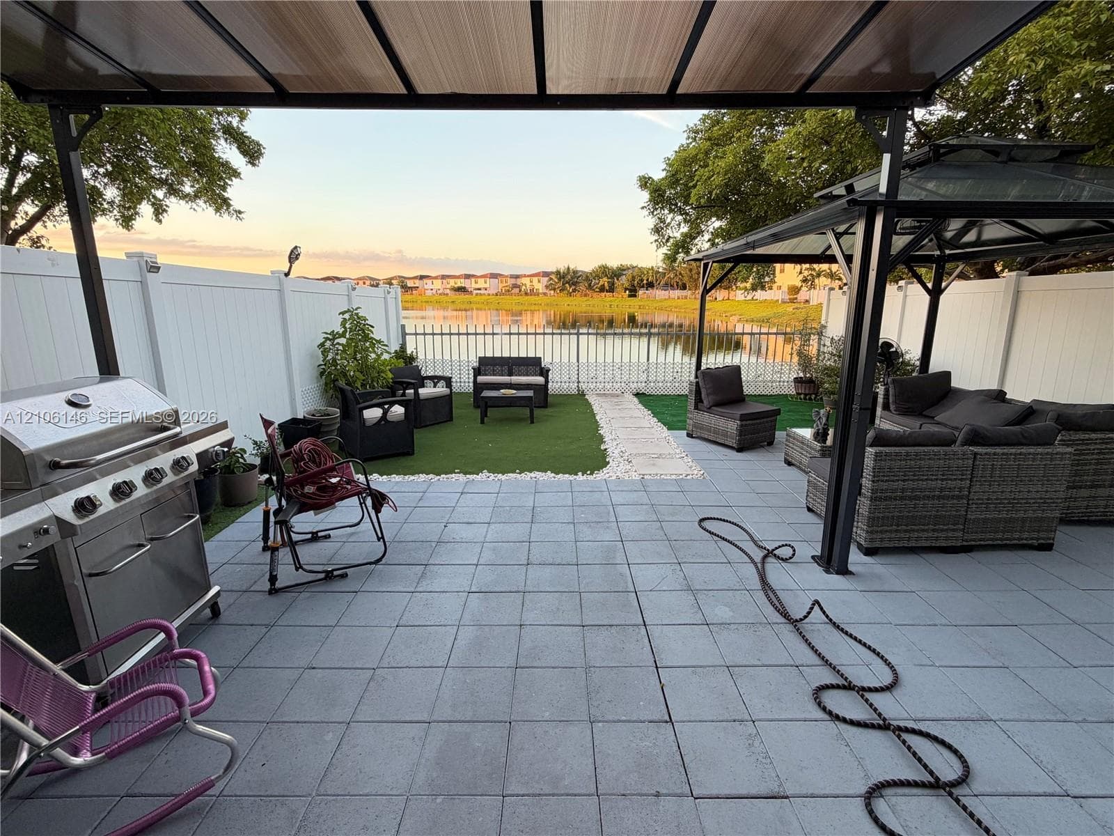 Paved patio featuring a metal gazebo, synthetic turf lawn, white vinyl fencing, and lake frontage
