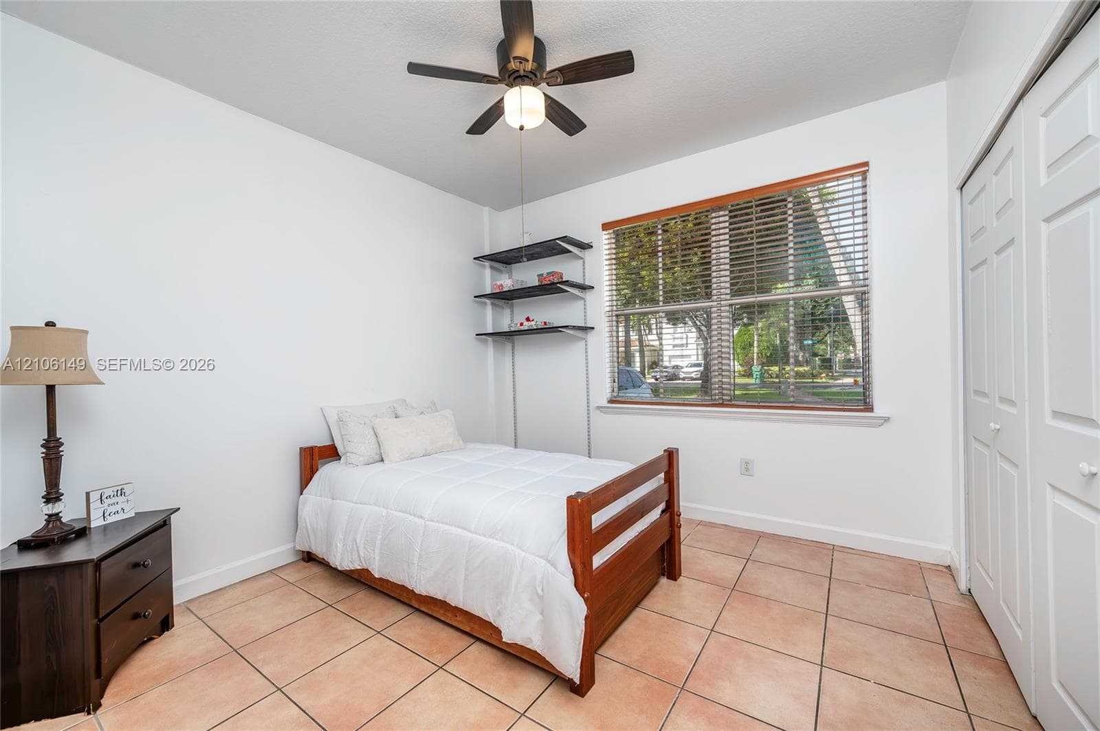 Bedroom featuring tile flooring, ceiling fan with integrated lighting, window with horizontal blinds, built-in shelving, and dual-panel closet doors