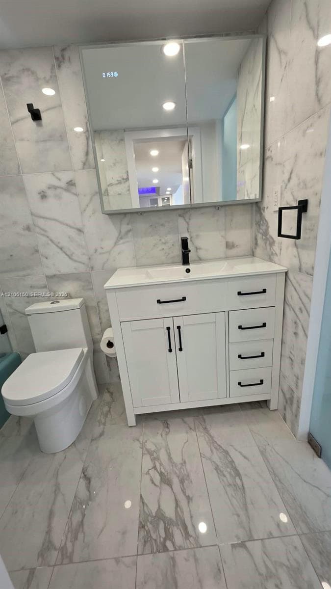 White vanity with matte black hardware, integrated sink, large medicine cabinet with integrated lighting, full-height stone-finish wall tiling, and matching stone-finish floor tile