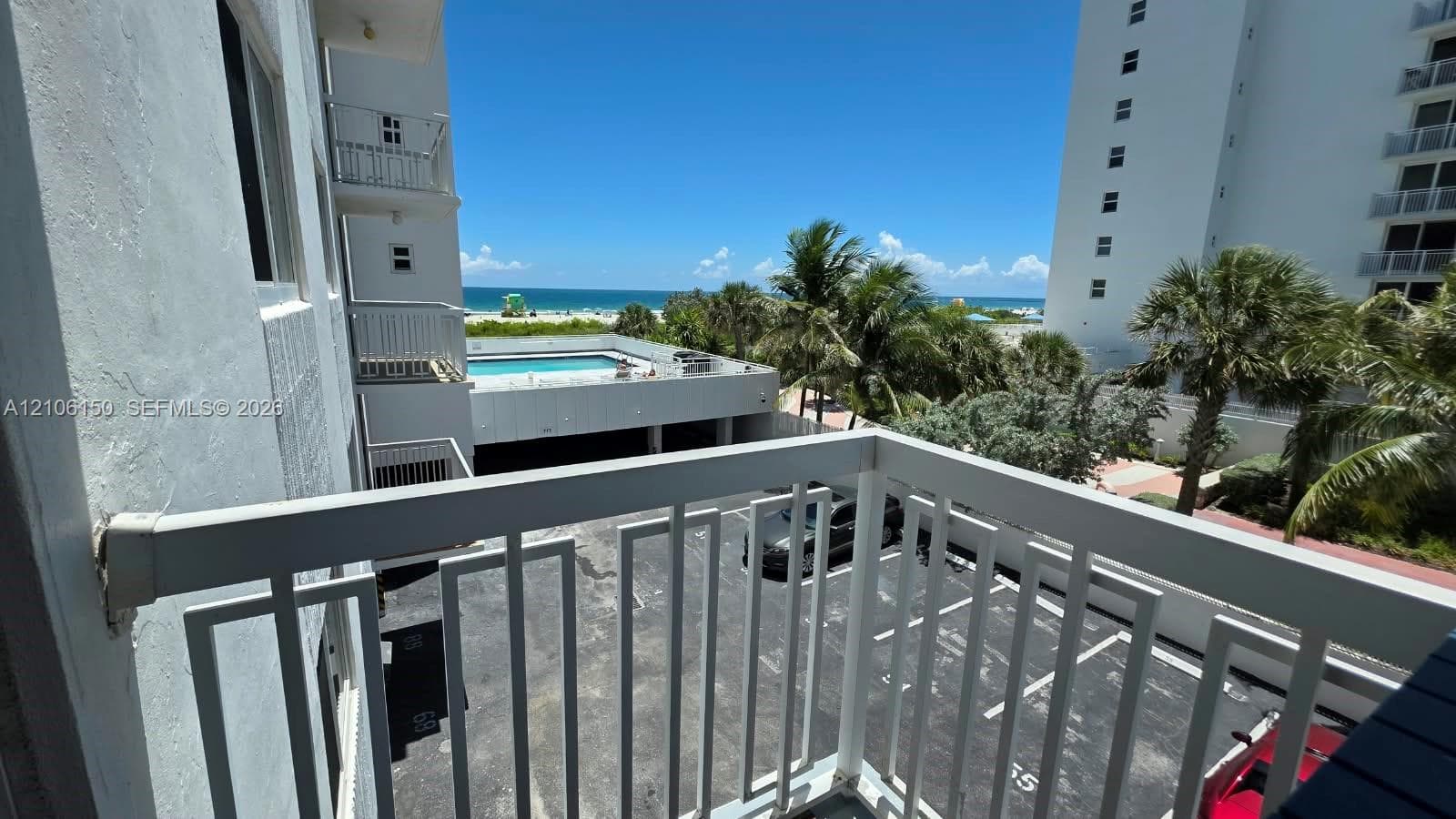 Balcony with metal railing overlooking the pool, parking area, and oceanfront horizon