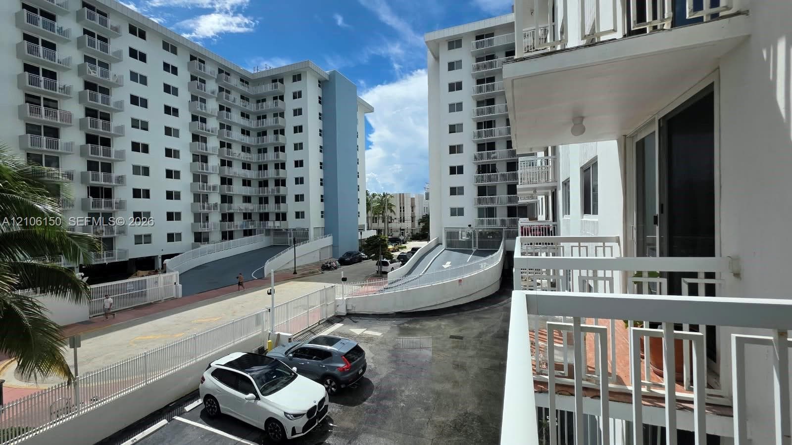 High-rise residential exterior featuring white stucco facade, multiple private balconies with metal railings, and integrated surface parking