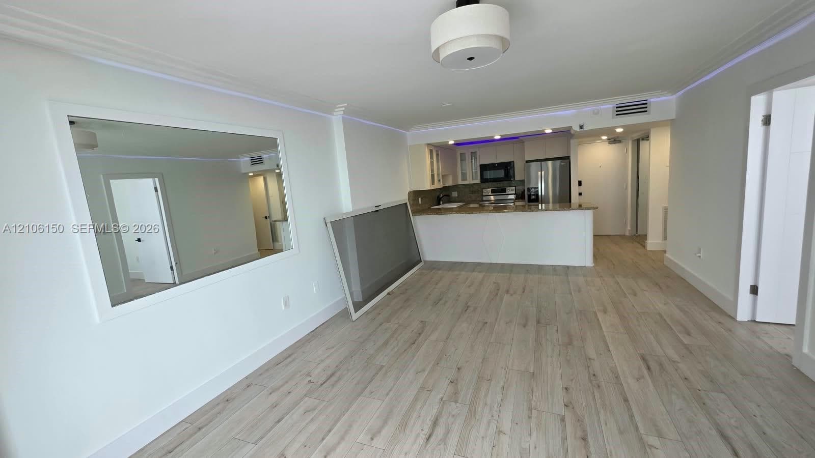 Open-concept interior featuring light wood-finish flooring, recessed crown molding with integrated perimeter accent lighting, a kitchen breakfast bar, and a wall-mounted framed mirror