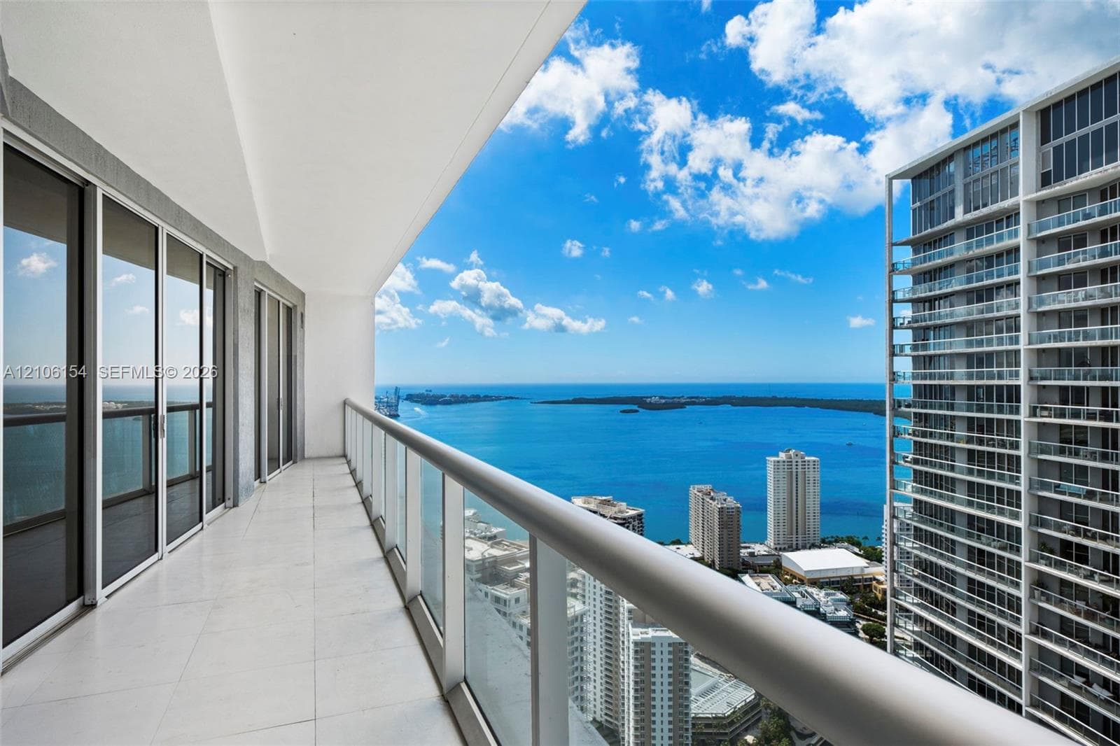 Expansive covered balcony featuring large-format tile flooring, a glass-panel railing with a minimalist handrail, and floor-to-ceiling sliding glass doors