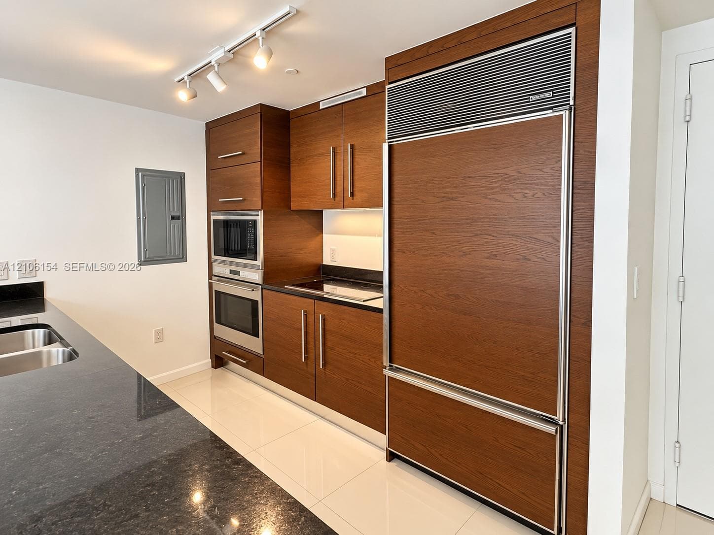 Kitchen featuring wood-finish cabinetry, panel-ready refrigerator, built-in stainless steel double oven and microwave, dark stone countertops, and ceiling-mounted track lighting