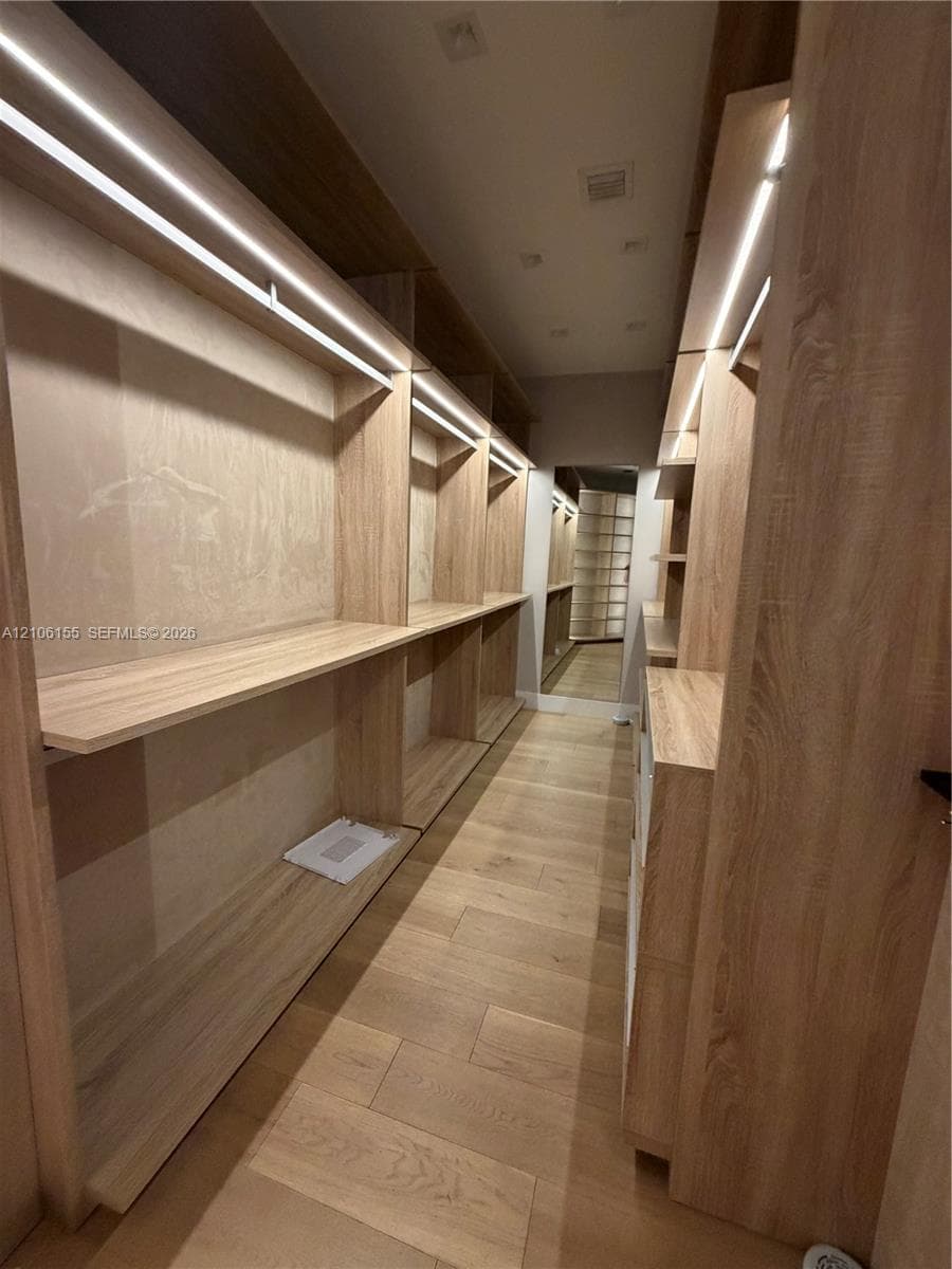 Custom Men’s Primary closet w storage system featuring light wood-finish cabinetry, integrated linear LED shelving illumination, open-concept shelving units, and wood-finish flooring