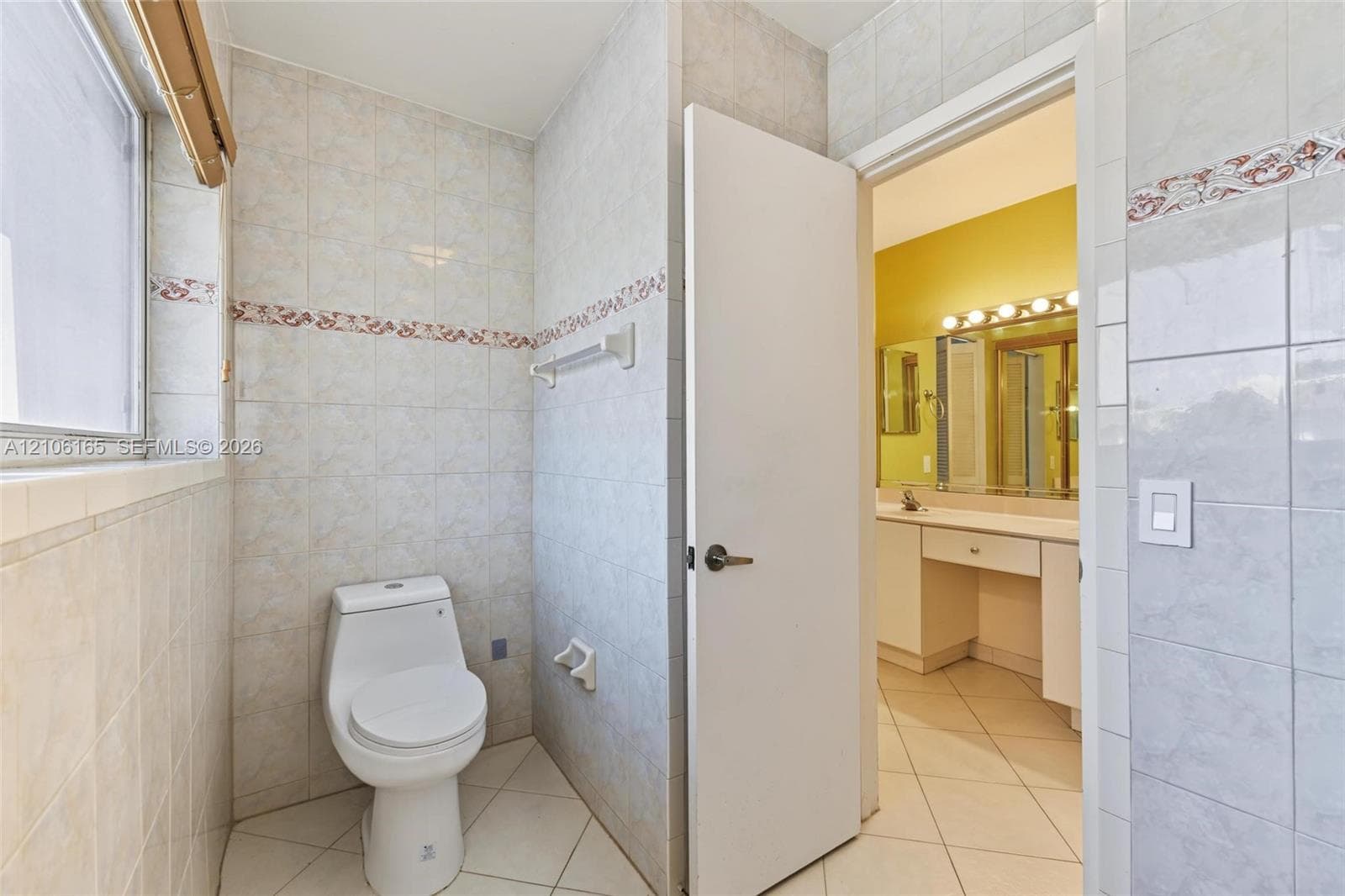 Bathroom featuring full-height ceramic tile walls with decorative border inlay, porcelain floor tile, white toilet, and built-in vanity with integrated lighting