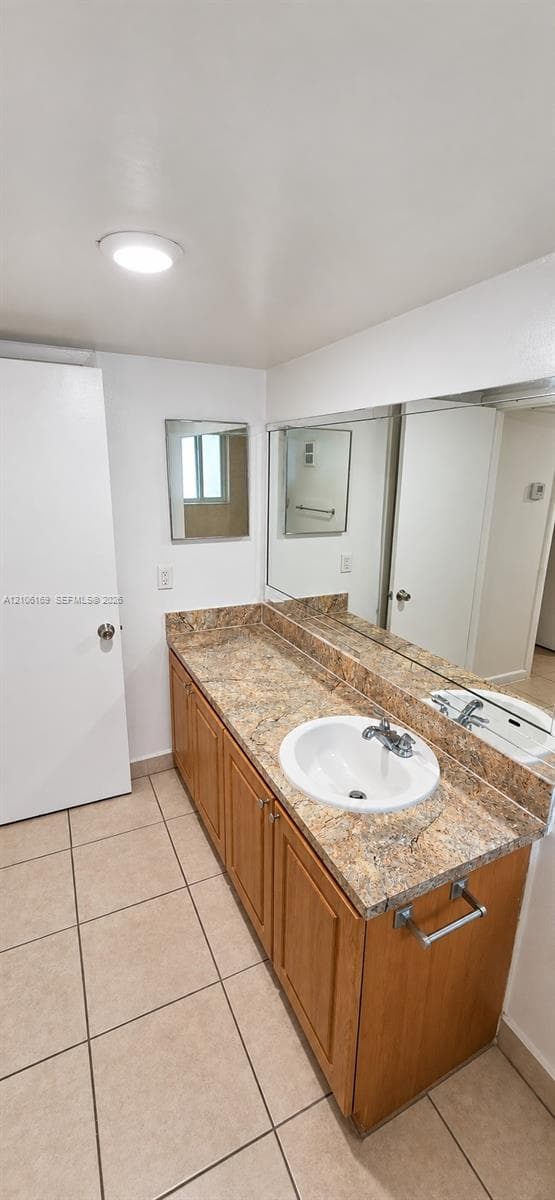 Vanity featuring wood-finish cabinetry, a stone-patterned countertop, an integrated circular sink, a full-width wall mirror, and neutral floor tiling