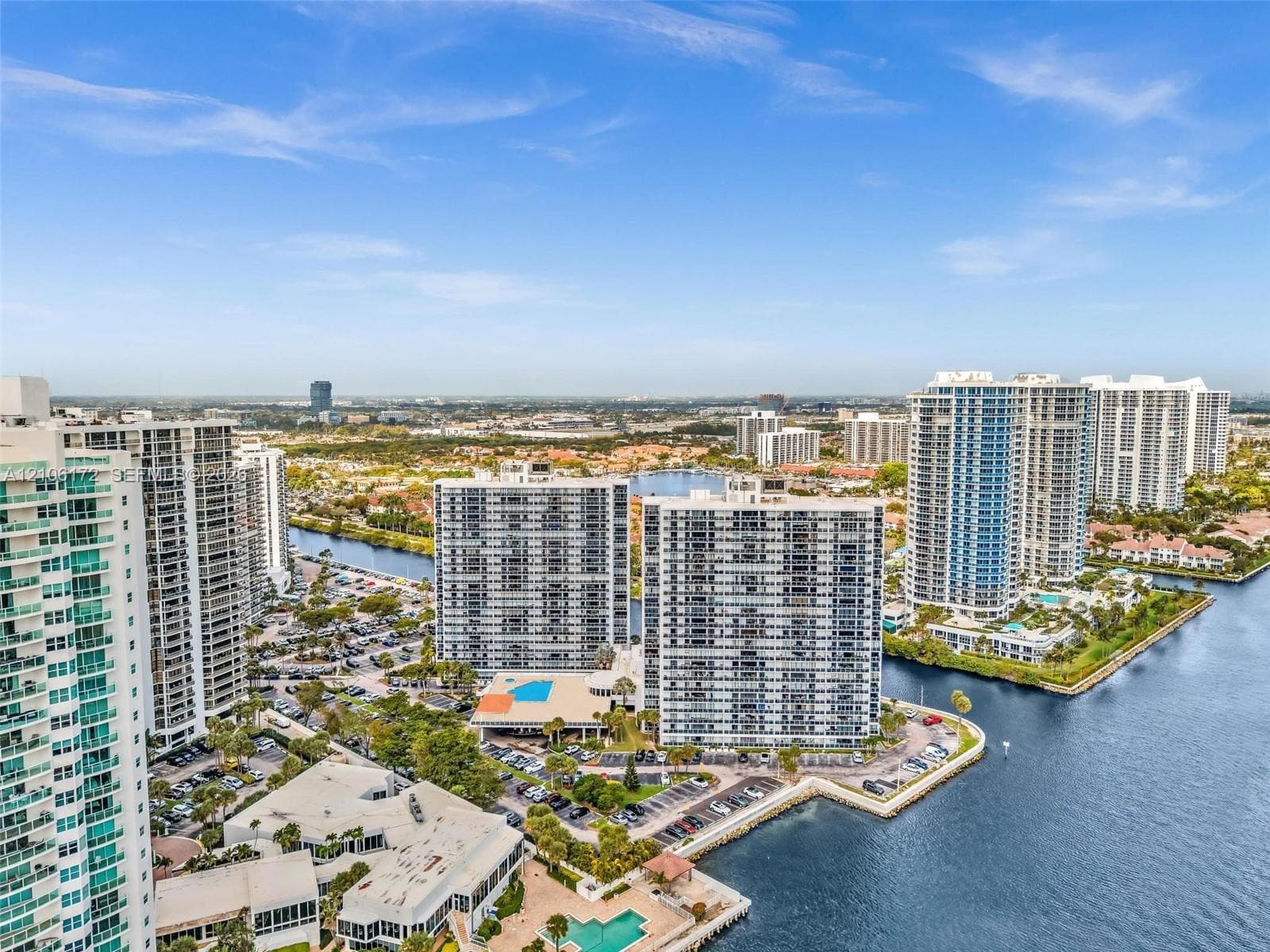 Aerial perspective of high-rise residential buildings situated along a waterway, featuring manicured landscaping, paved parking areas, and ground-level swimming pools