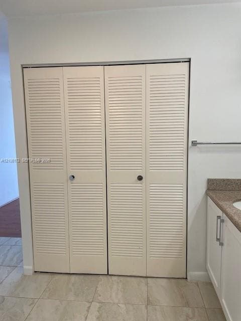 Bi-fold closet doors featuring white louvered panels, light-toned stone-finish tile flooring, and built-in vanity with a stone-finish countertop