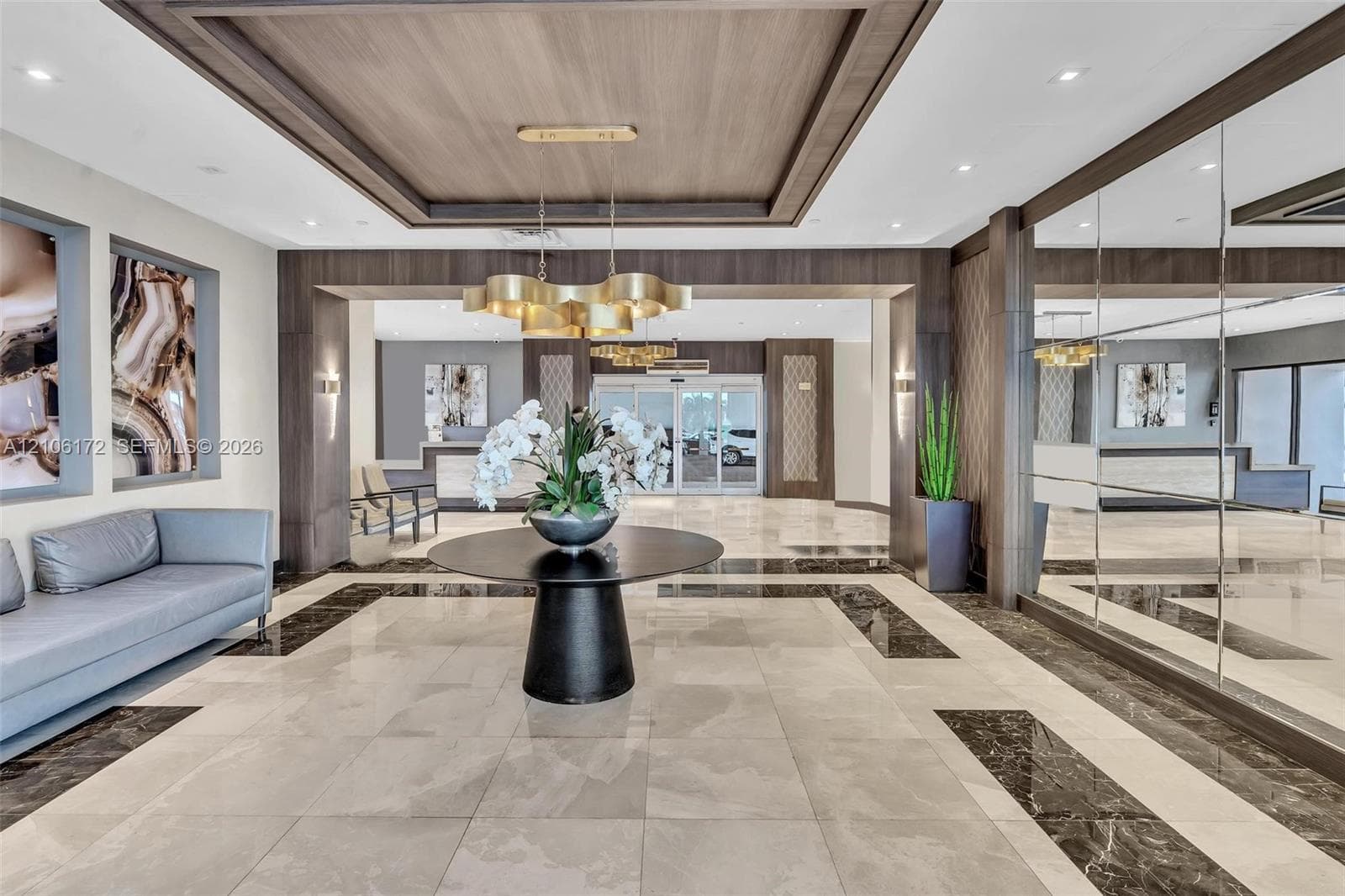 Grand lobby featuring tile flooring with dark stone inlay, coffered wood-paneled ceiling, large-scale gold-finish chandelier, and full-height mirrored wall panels