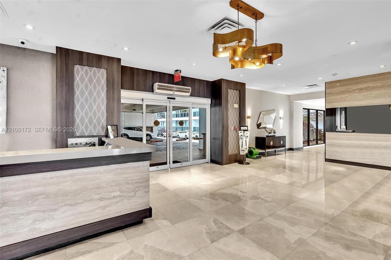 Lobby area featuring polished tile flooring, a custom reception desk with stone-finish surfaces and dark wood-tone cabinetry, a sculptural gold-tone pendant light fixture, and floor-to-ceiling glass entry doors