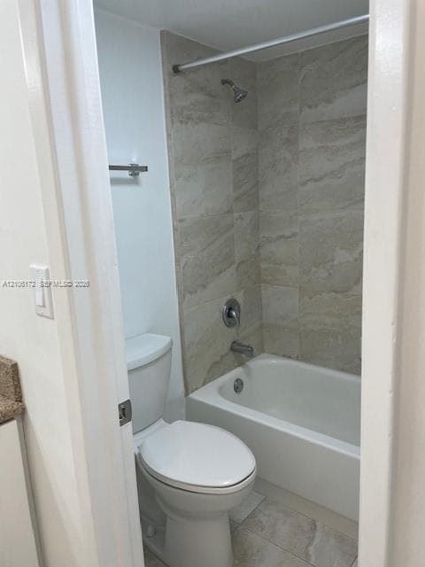 Bathroom featuring a white porcelain tub with integrated shower, stone-finish tile surround, floor-mounted toilet, and wall-mounted towel bar