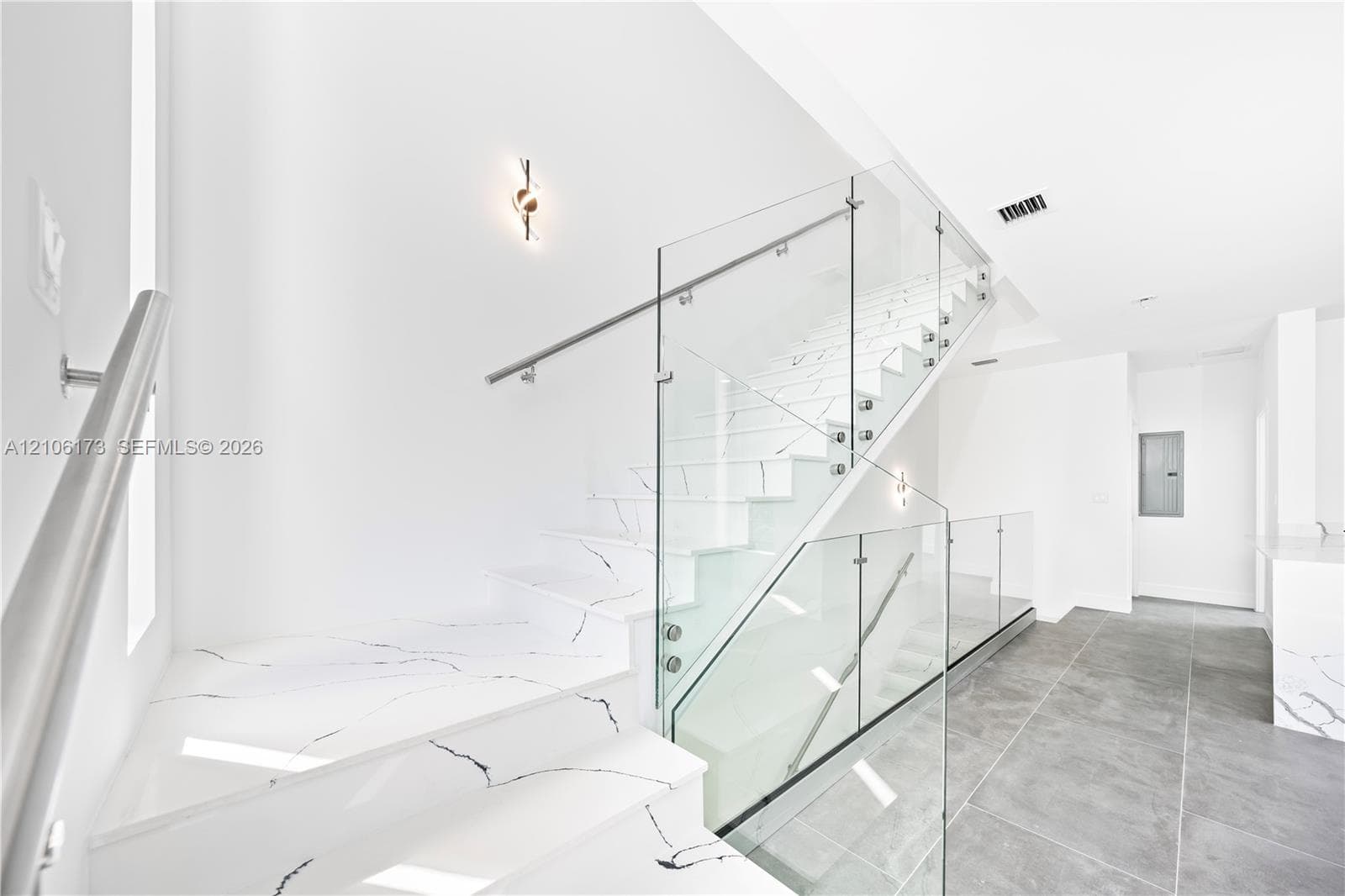 Staircase featuring white treads with veined pattern, frameless glass panel railing, and brushed metal wall-mounted handrail, complemented by large-format grey tile flooring