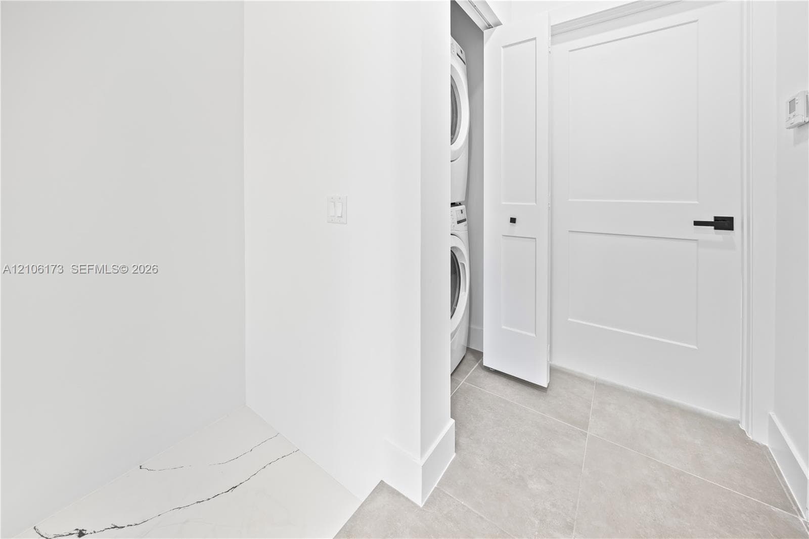 Laundry area featuring a stacked washer and dryer, large-format tile flooring, a solid-core door with matte black hardware, and stone-finish built-in counter surface
