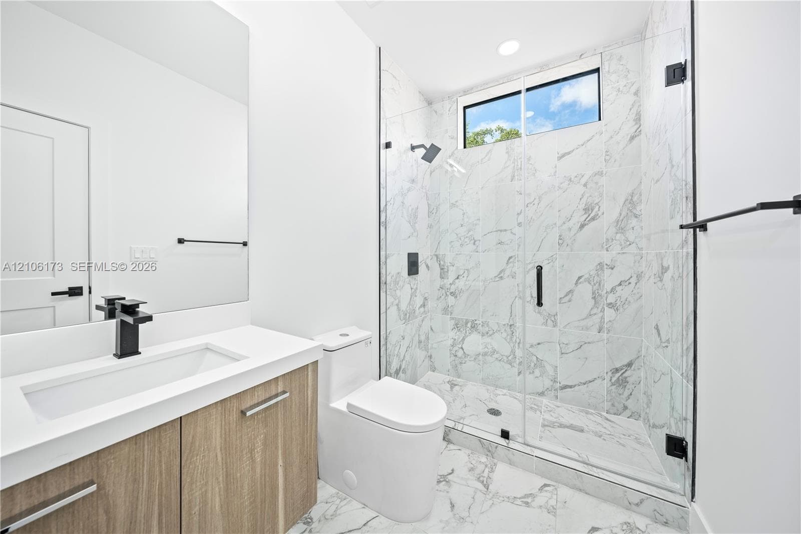 Floating vanity with integrated sink, stone-patterned wall and floor tile, glass-enclosed shower, transom window, and matte black fixtures