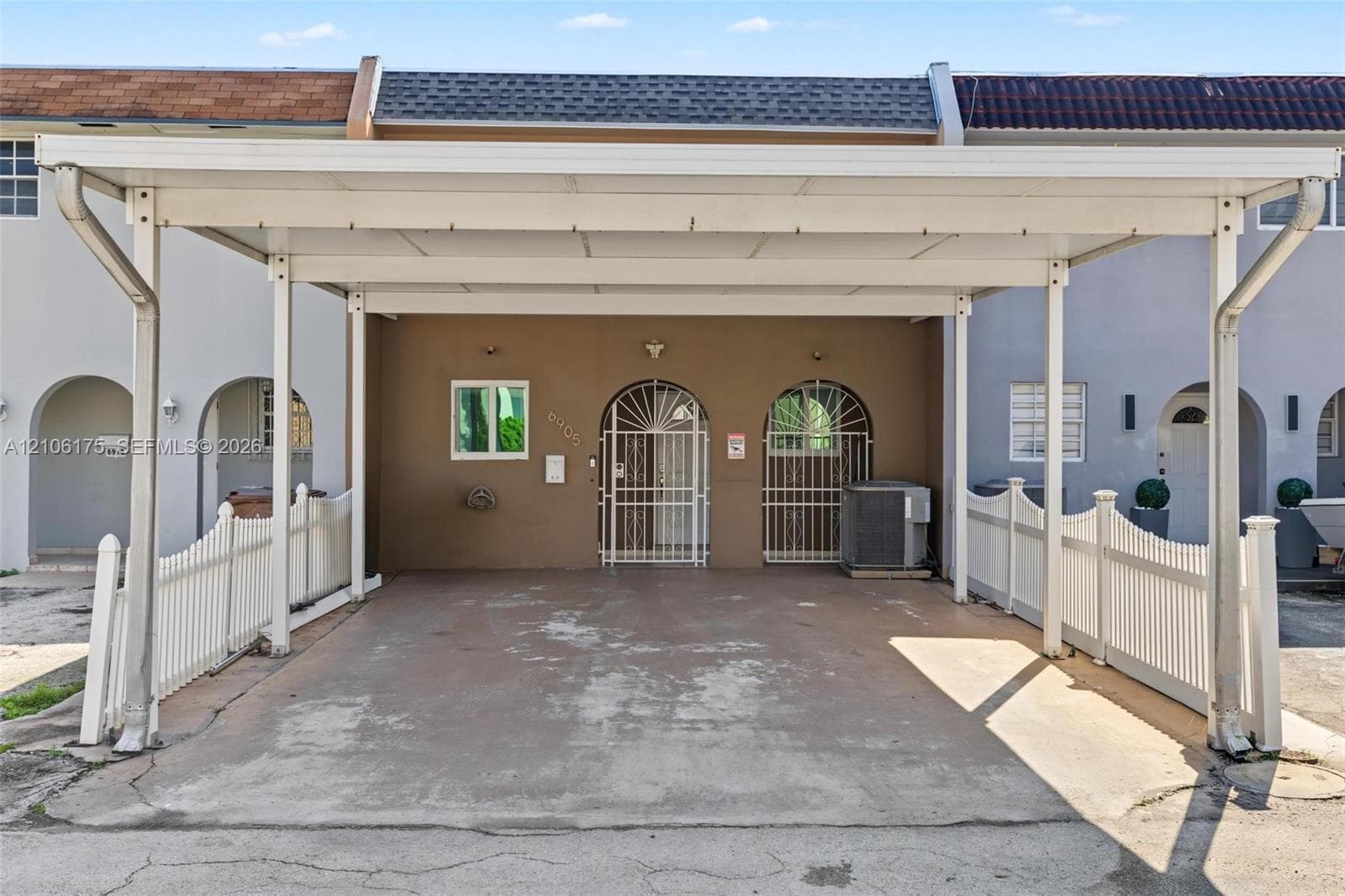 Covered carport featuring a concrete parking pad, white vinyl perimeter fencing, and dual arched metal security doors
