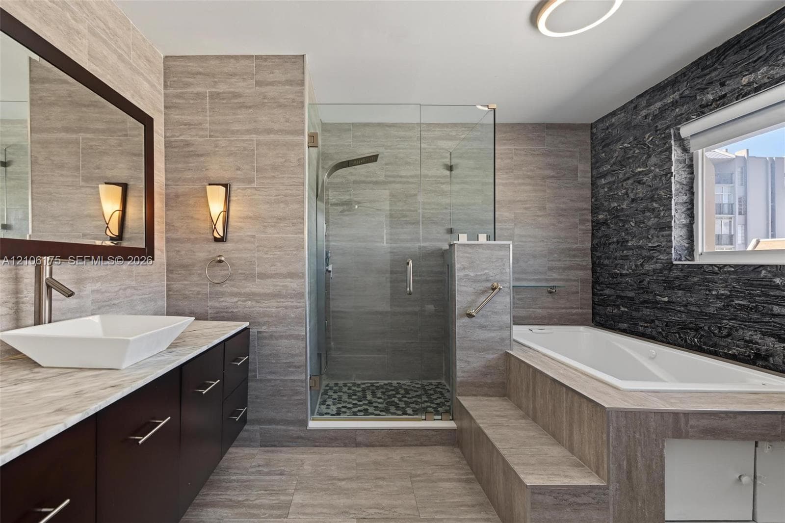 Bathroom featuring a floating vanity with a rectangular vessel sink, glass-enclosed shower with mosaic tile flooring, built-in soaking tub with a stepped surround, and dark stone accent wall