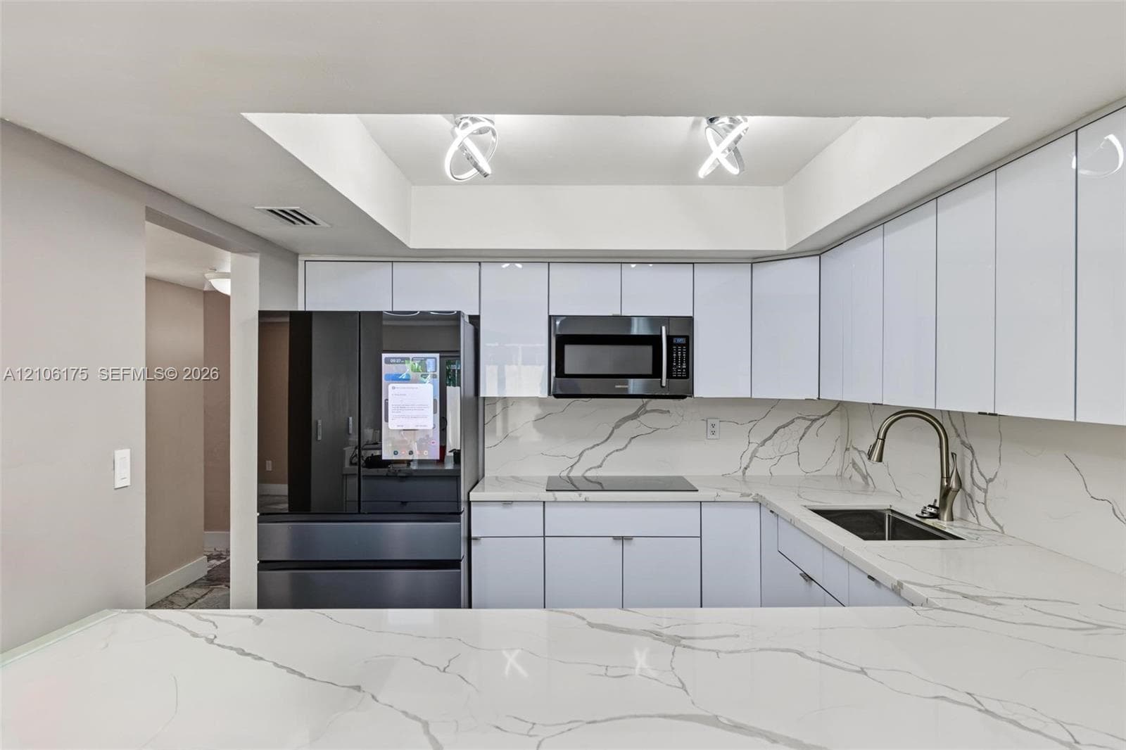 Kitchen featuring high-gloss white cabinetry, stone-patterned countertops, a matching full-height backsplash, a stainless steel undermount sink with a gooseneck faucet, and recessed tray ceiling lighting