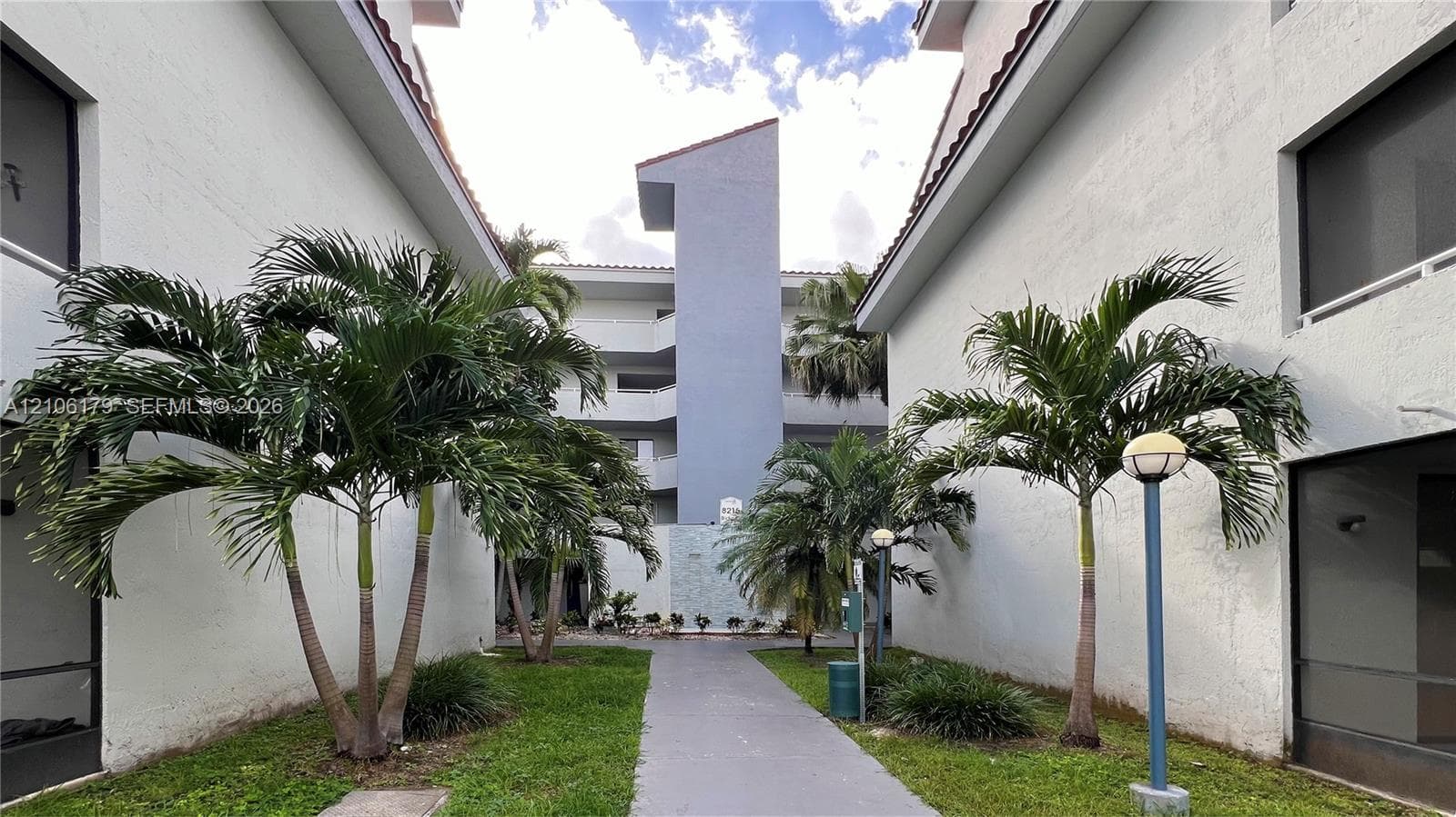 Multi-story stucco exterior featuring a paved walkway, mature palm trees, manicured landscaping, and post-mounted outdoor lighting