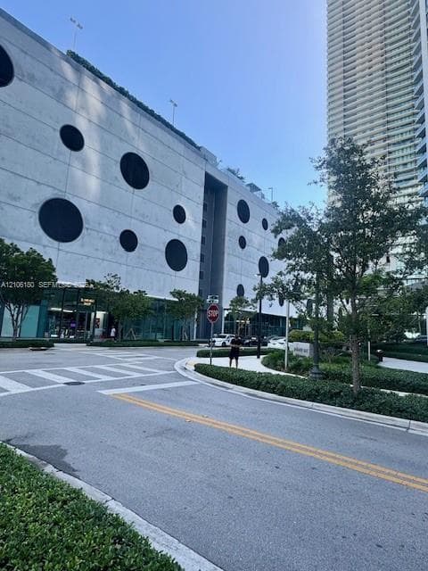 Multi-level concrete structure featuring a geometric circular facade, ground-level glass storefronts, and landscaped perimeter hedging
