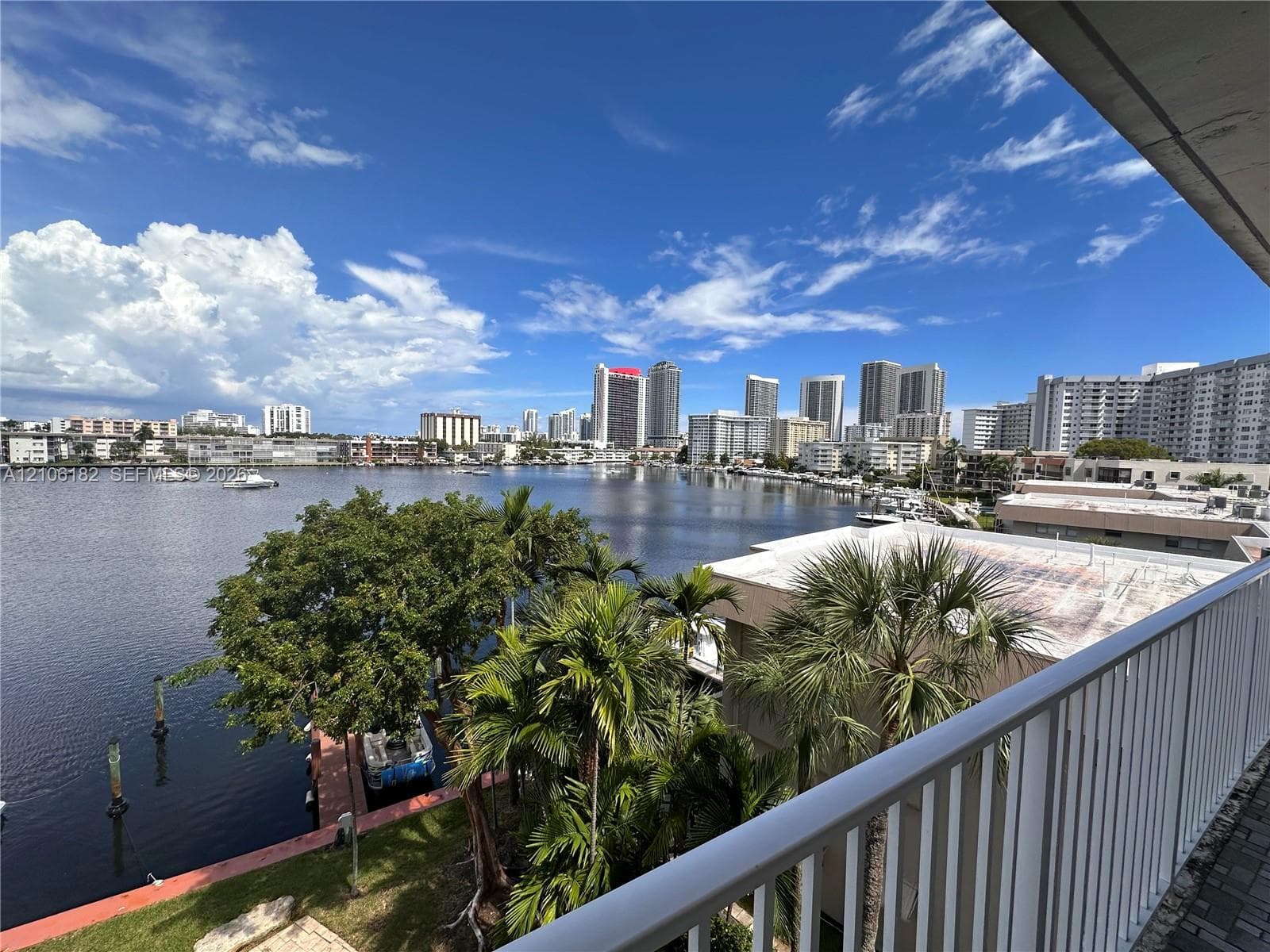 Elevated balcony featuring a white metal railing, overlooking a waterway with mature palm trees and a skyline backdrop