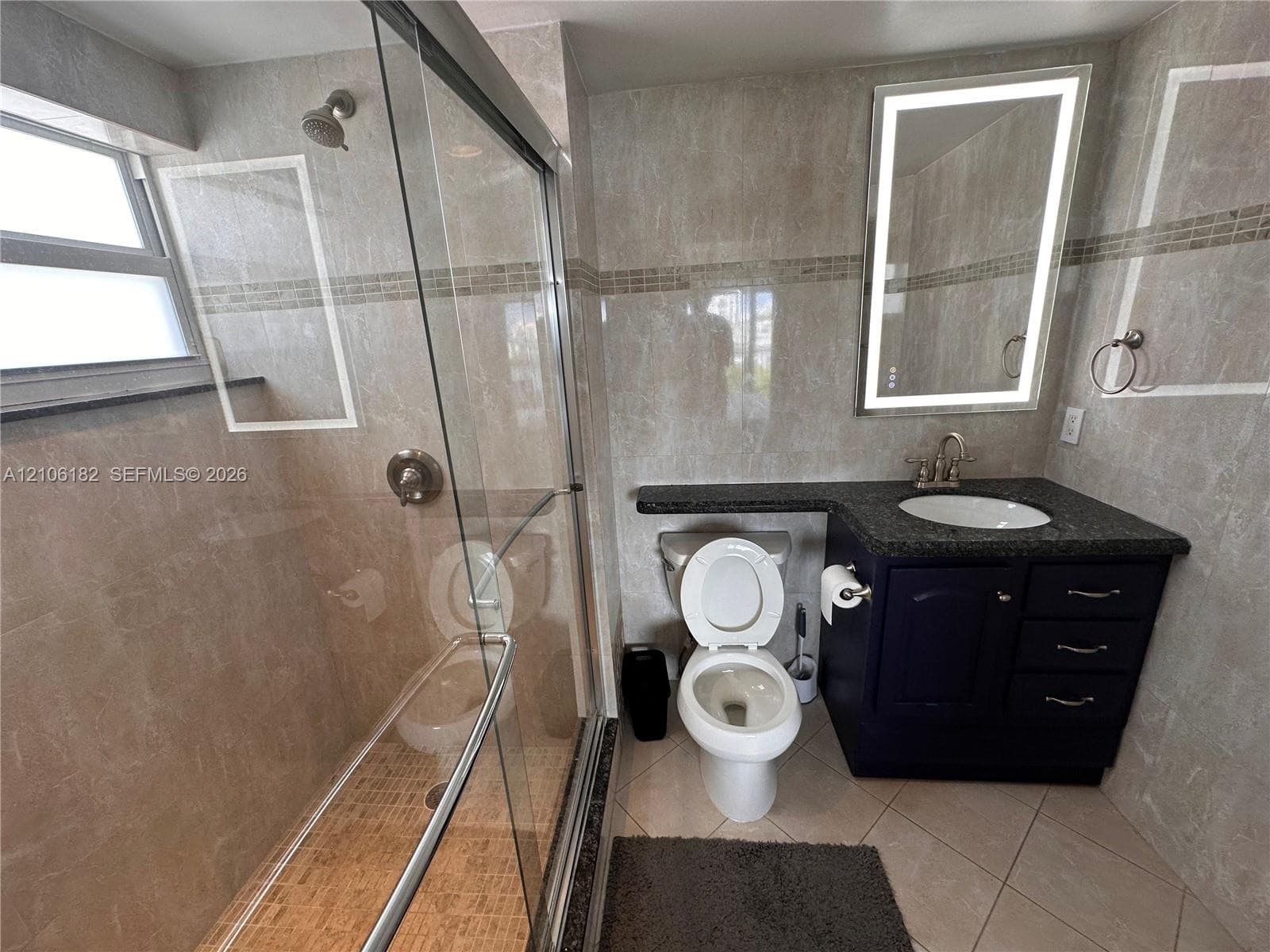 Walk-in shower with glass enclosure, dark cabinetry vanity with integrated countertop, oval undermount sink, and illuminated mirror