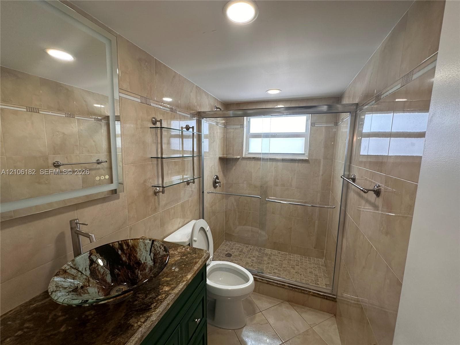 Bathroom featuring tile wall surfaces, a vessel sink with a stone-finish countertop, a walk-in shower with a glass enclosure, and a wall-mounted light-up mirror