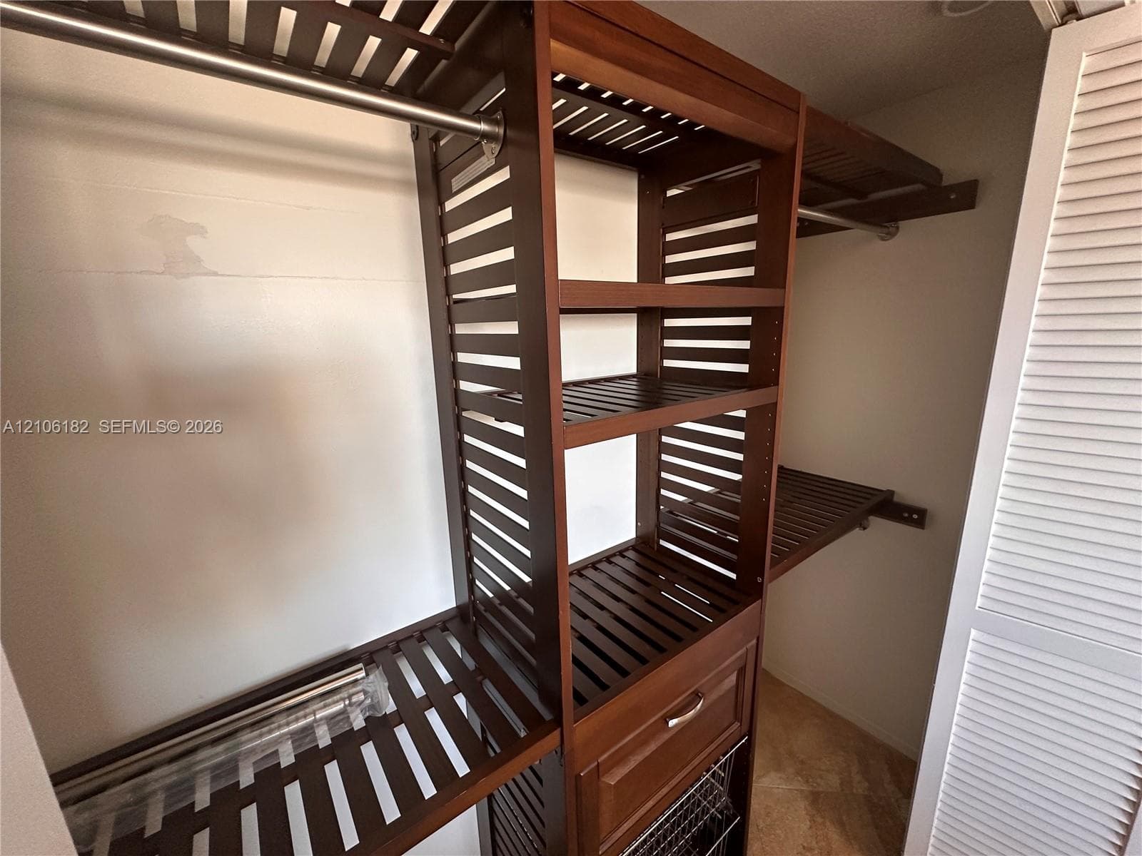 Custom closet system featuring dark wood-finish open shelving, integrated hanging rods, a built-in drawer, and a lower pull-out wire basket, secured behind louvered doors