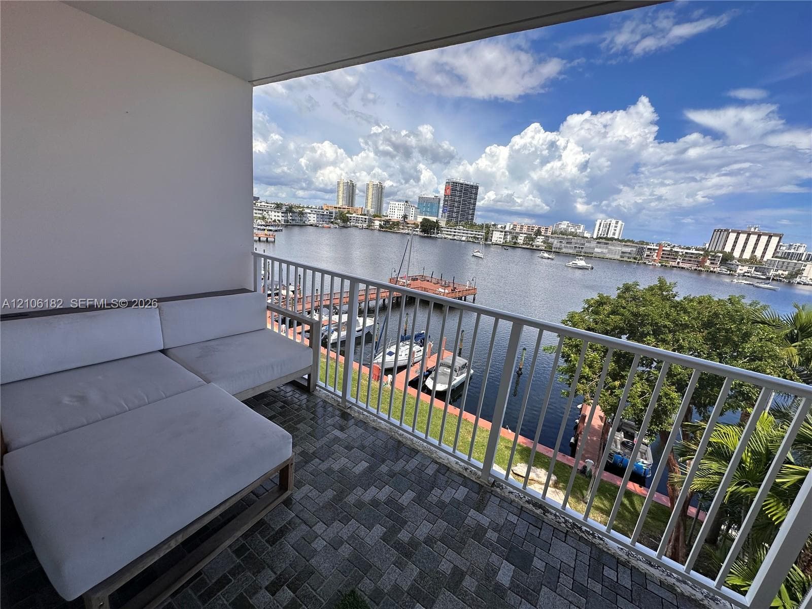 Covered balcony featuring patterned stone paver flooring and a white metal safety railing overlooking a deep-water canal, boat slips, and a cityscape
