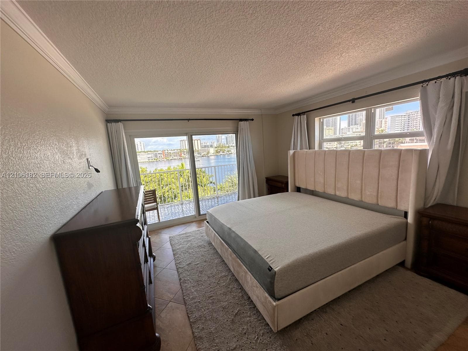 Floor-to-ceiling sliding glass doors, neutral tile flooring, crown molding, and a private balcony featuring a water backdrop