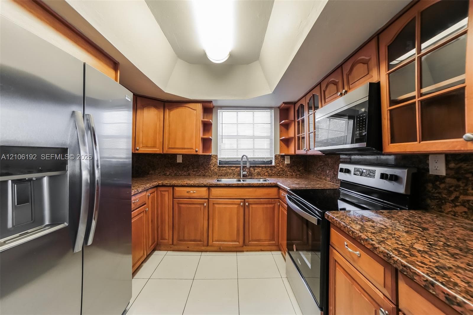Kitchen featuring warm-tone cabinetry, stone-finish countertops, stainless steel appliances, tile flooring, and a tray ceiling with recessed lighting