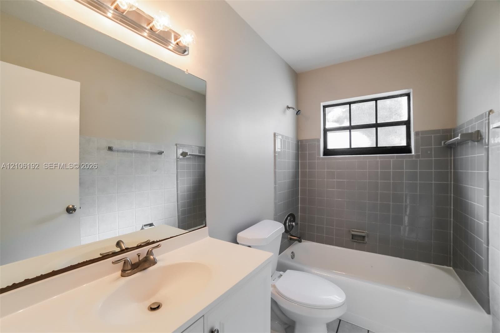 Bathroom featuring a vanity with integrated sink, expansive wall-mounted mirror, multi-bulb light fixture, and a tub-shower combination with gray tile surround and window