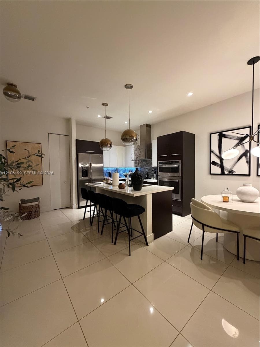 Modern kitchen featuring flat-panel cabinetry, a central island with stone-surface countertop, stainless steel double wall oven, stainless steel refrigerator, and large-format tile flooring