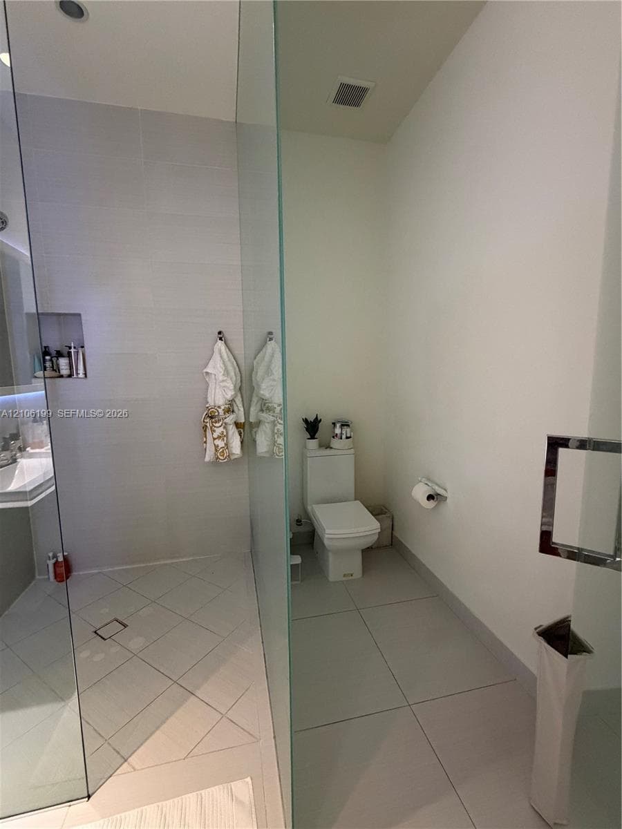 Walk-in shower featuring large-format tile flooring, wall-mounted shower niche, frameless glass enclosure, and white porcelain toilet