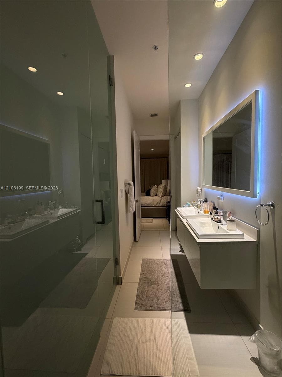 Floating dual-sink vanity with a rectangular mirror featuring integrated LED backlighting, glass-enclosed shower, recessed ceiling lighting, and large-format tile flooring
