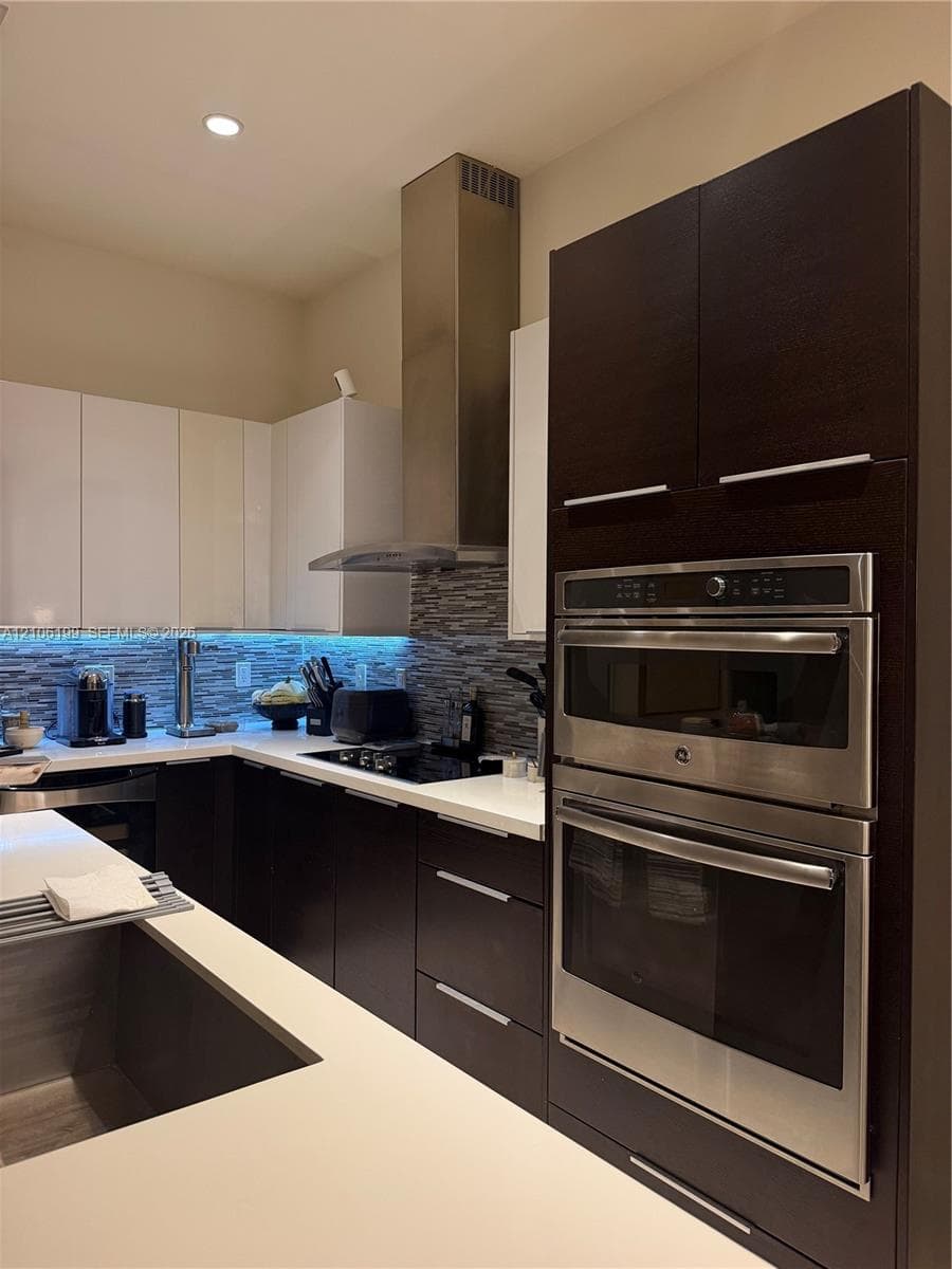 Kitchen featuring high-contrast cabinetry, stone countertops, stacked-tile backsplash, stainless steel built-in wall oven and microwave, and a professional-grade stainless steel range hood