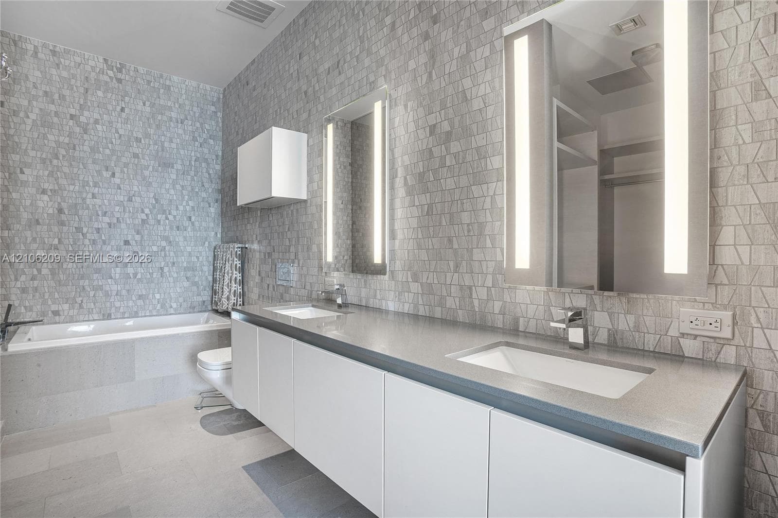 Double vanity with solid surface countertops, integrated rectangular sinks, wall-mounted backlit mirrors, textured tile wall cladding, and a deep soaking tub