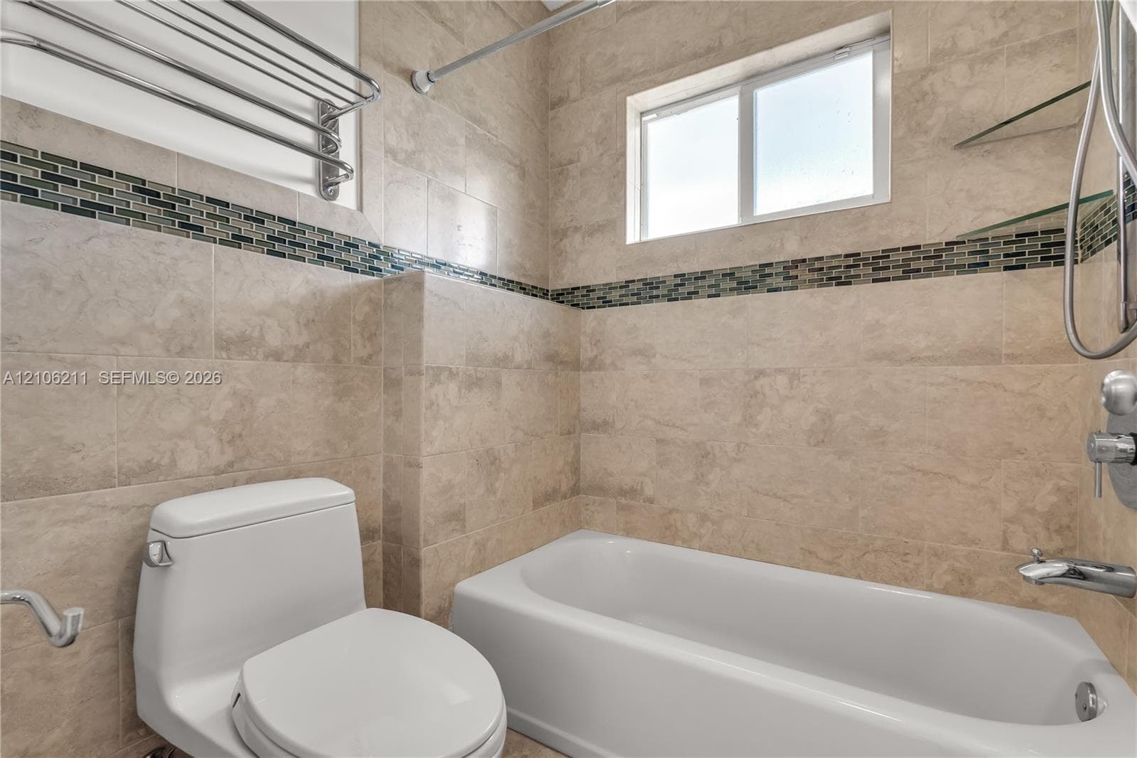 Full-height wall tiling featuring natural stone-finish tiles and a decorative glass mosaic accent band, white bathtub with chrome plumbing fixtures, integrated glass shelving, and a wall-mounted metal towel rack