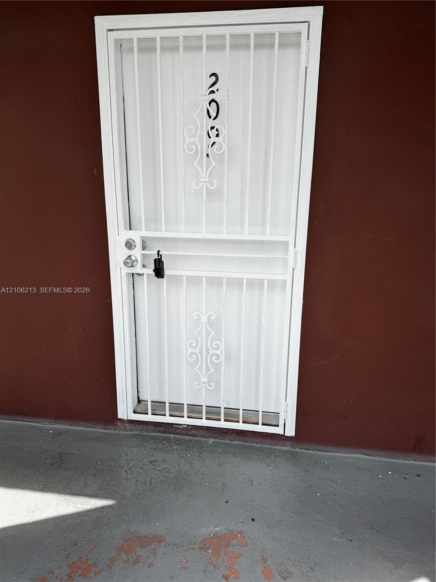 White metal security screen door featuring decorative scrollwork, a vertical bar pattern, and a dual-locking mechanism