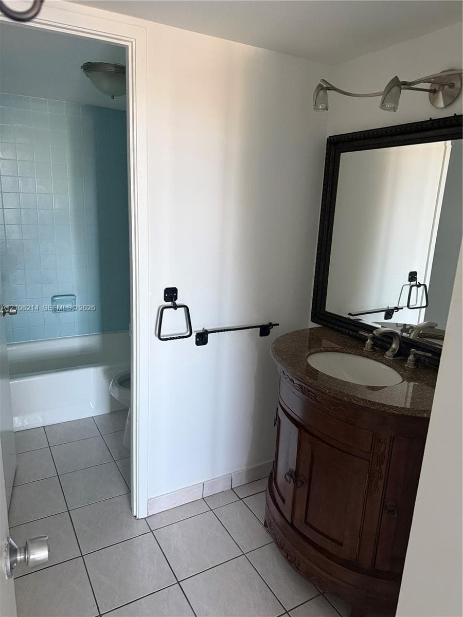 Wood-finish vanity with a stone countertop, integrated undermount sink, multi-light fixture, framed wall mirror, and tile flooring