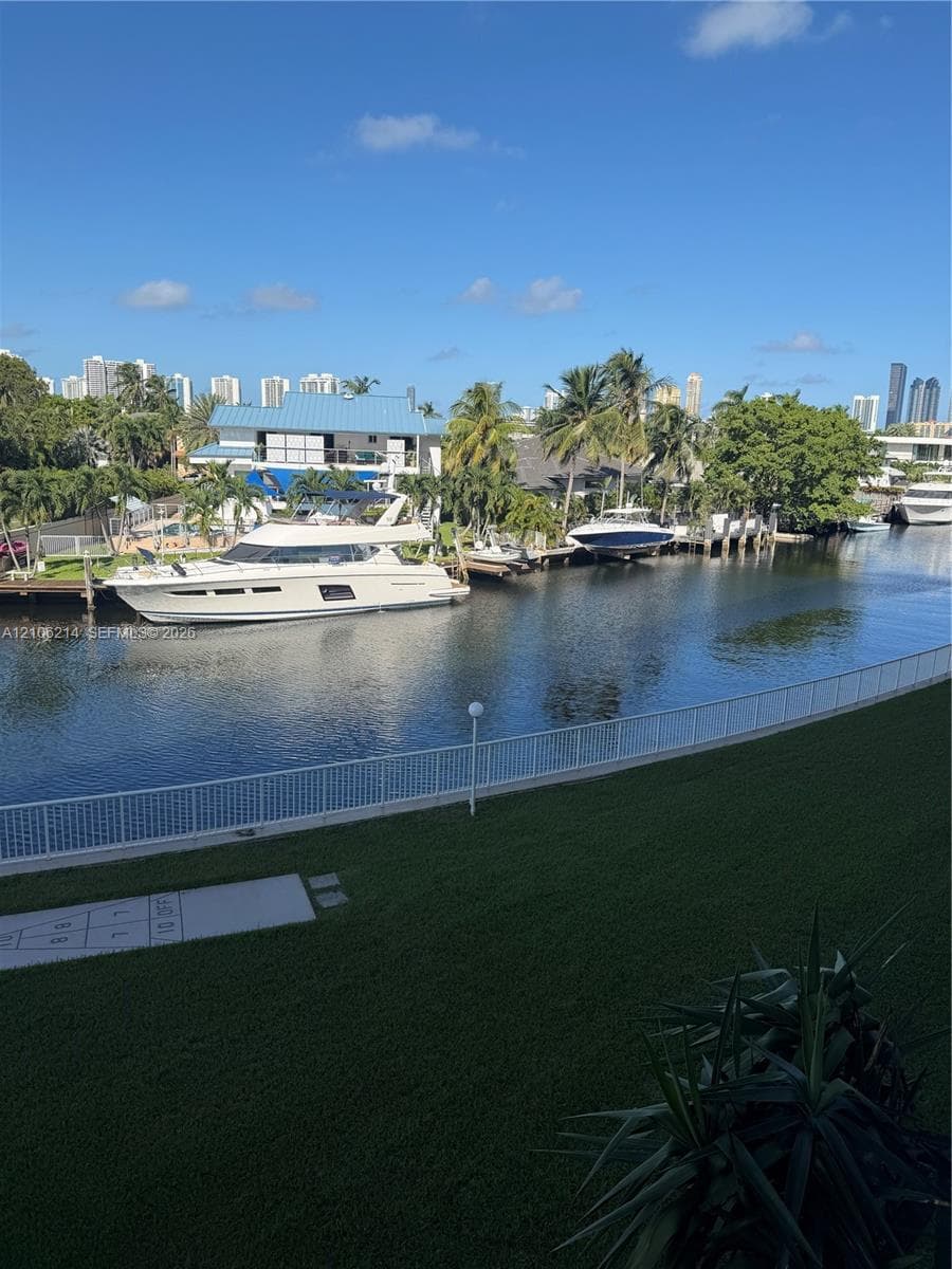Waterfront property featuring a canal bulkhead, manicured lawn, white metal fencing, and a concrete hopscotch court