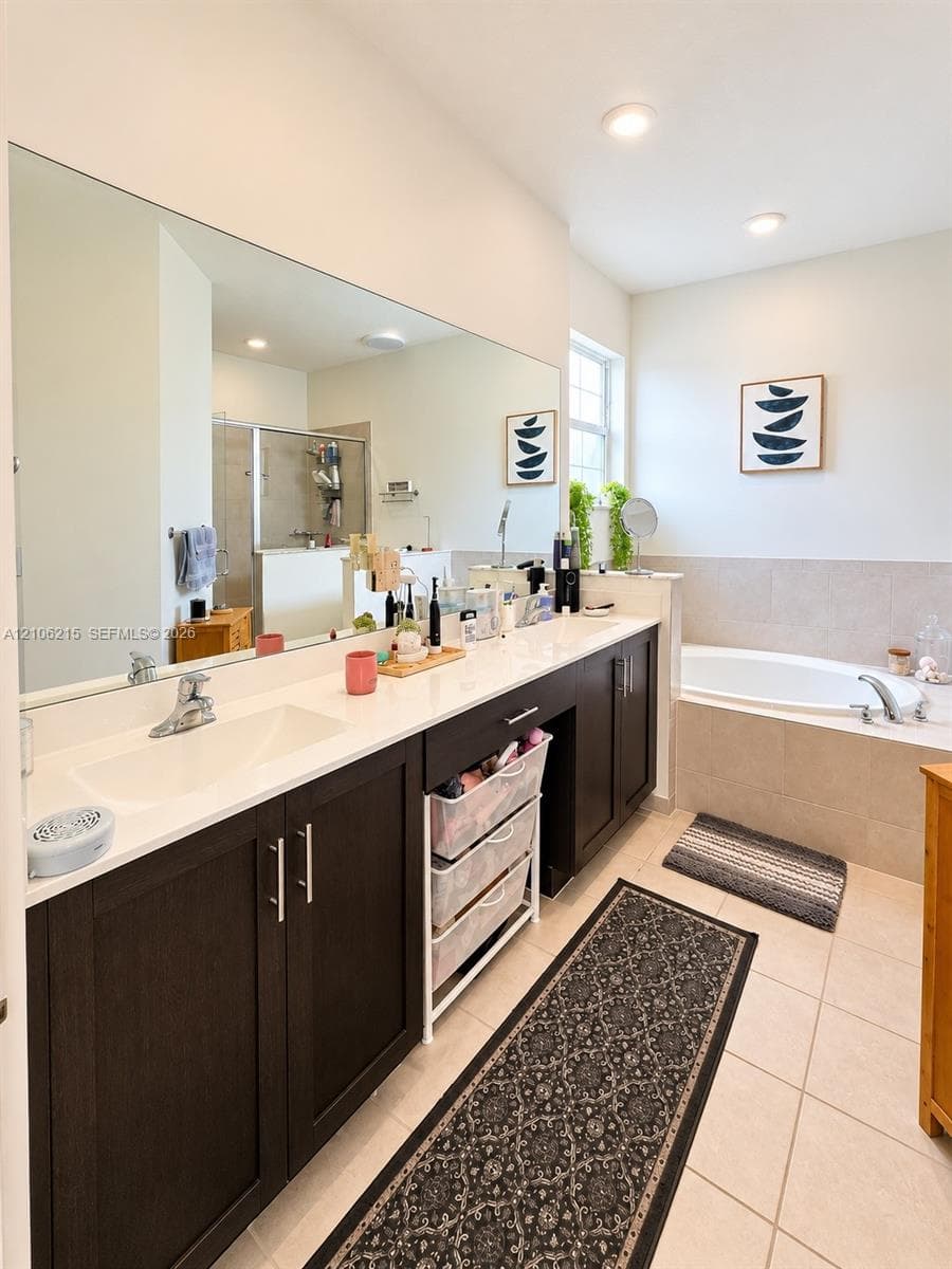 Bathroom featuring a dual-sink vanity with dark cabinetry, stone-finish countertops, full-width mirror, tile flooring, and a deep soaking tub