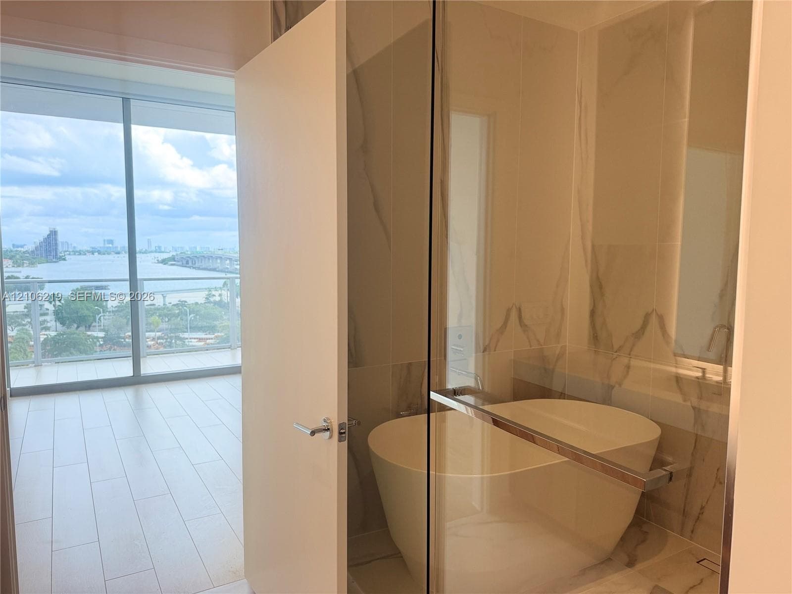 Freestanding soaking tub with a glass partition, large-format stone-finish tile surround, and floor-to-ceiling windows with water backdrop
