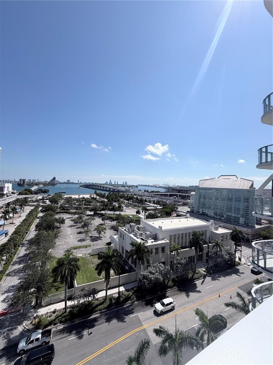 Elevated exterior perspective featuring glass-railed balconies, palm tree-lined streets, and panoramic water and bridge scenery