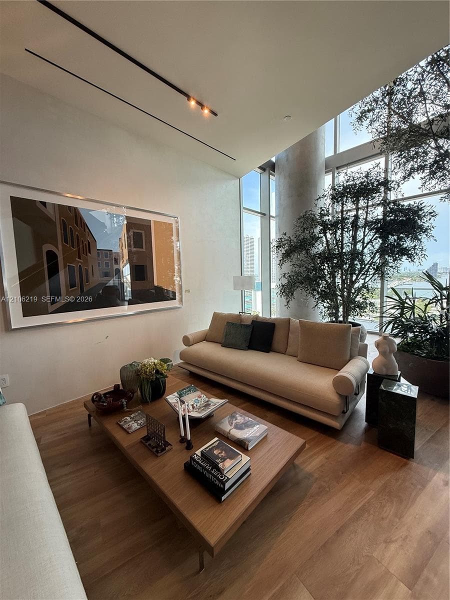 Double-height floor-to-ceiling windows, wood-finish flooring, recessed track lighting, and a prominent concrete structural column