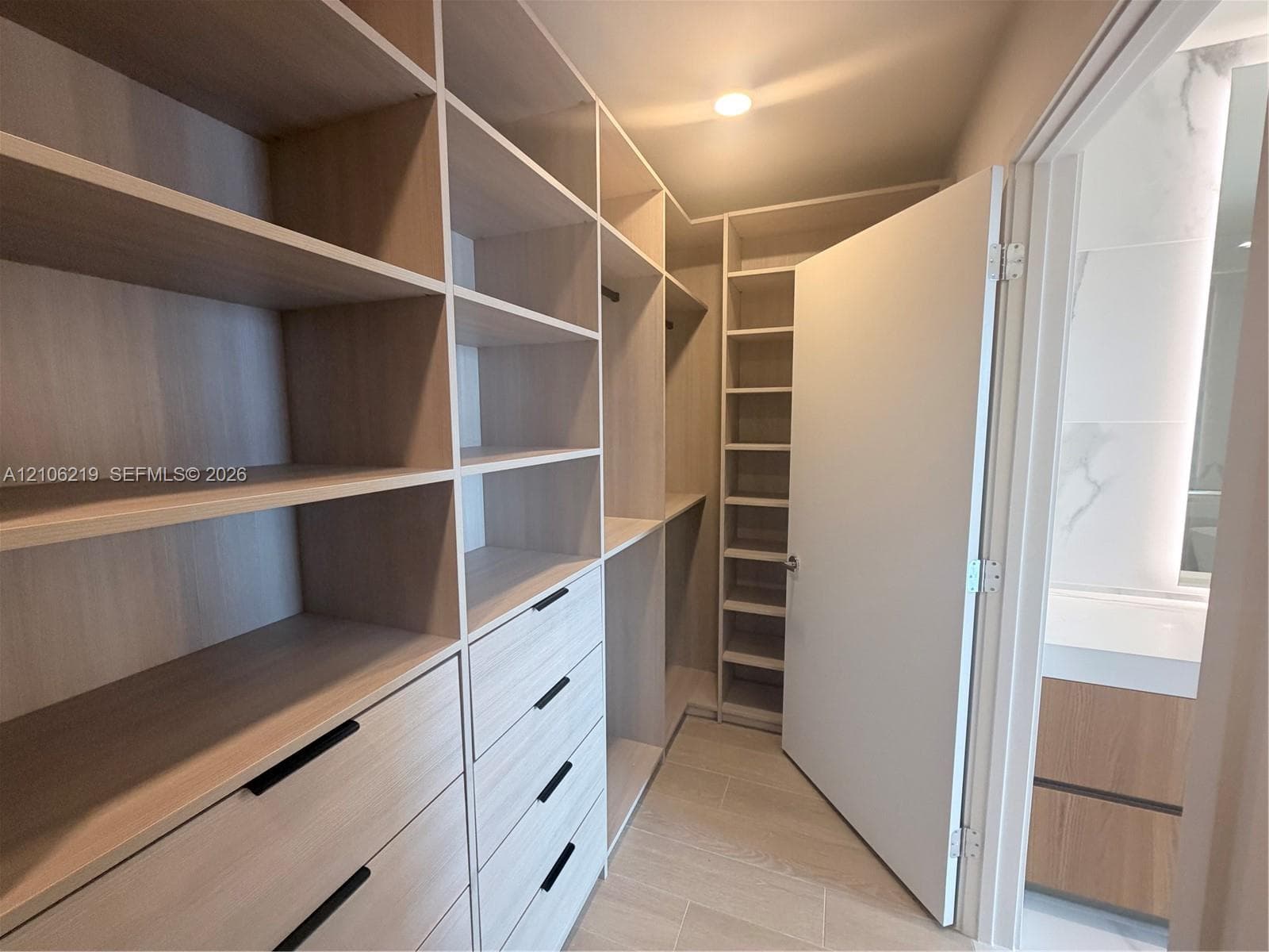 Walk-in closet featuring light-toned wood-finish built-in shelving, custom cabinetry with matte black hardware, recessed ceiling lighting, and large-format neutral-tone tile flooring